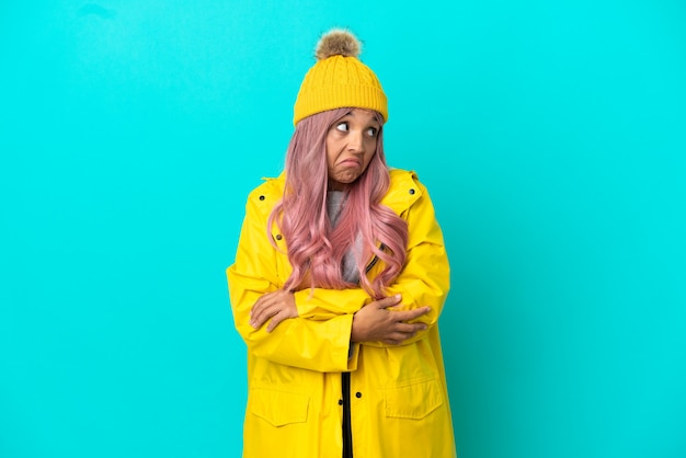 Photo jeune femme aux cheveux roses portant un manteau imperméable isolé sur fond bleu faisant un geste de doute tout en soulevant les épaules