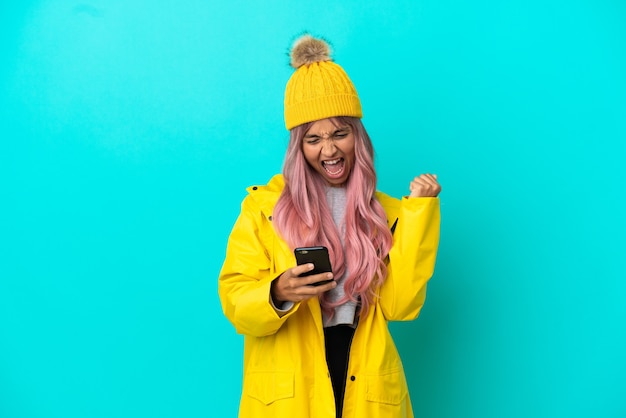 Jeune femme aux cheveux roses portant un manteau imperméable isolé sur fond bleu avec téléphone en position de victoire