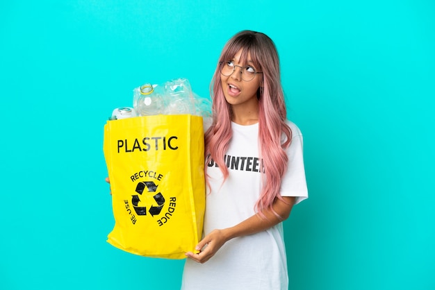 Jeune femme aux cheveux roses tenant un sac plein de bouteilles en plastique à recycler isolé sur fond bleu en pensant à une idée tout en levant les yeux