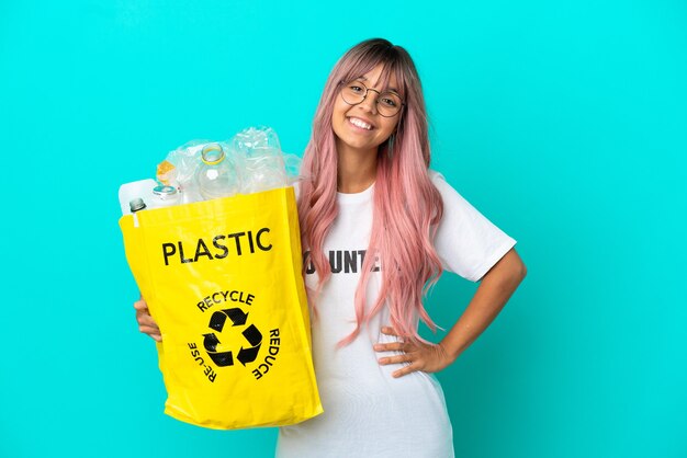 Jeune femme aux cheveux roses tenant un sac plein de bouteilles en plastique à recycler isolé sur fond bleu posant avec les bras à la hanche et souriant