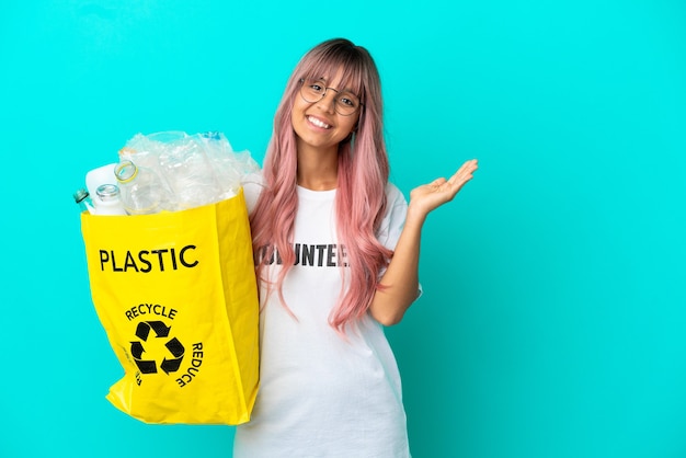 Jeune femme aux cheveux roses tenant un sac plein de bouteilles en plastique à recycler isolé sur fond bleu tendant les mains sur le côté pour inviter à venir