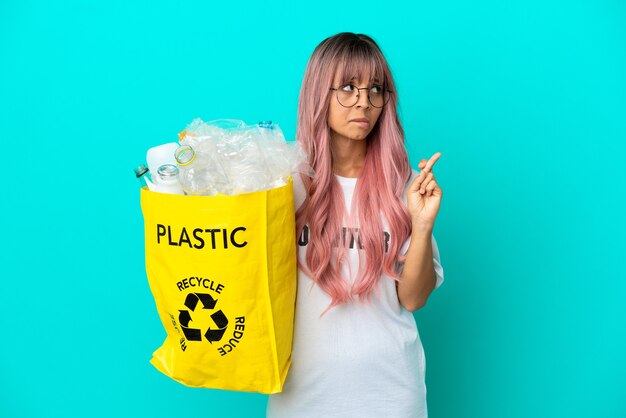 Jeune femme aux cheveux roses tenant un sac plein de bouteilles en plastique à recycler isolée sur fond bleu avec les doigts croisés et souhaitant le meilleur