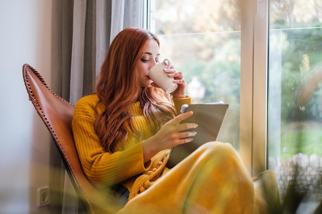 Jeune femme aux cheveux rouges lisant sur la tablette et buvant un café chaud près de la fenêtre à la maison