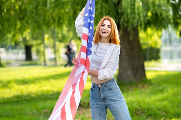 Jeune femme aux cheveux rouges tenant le drapeau national des USA debout à l'extérieur dans le parc d'été.