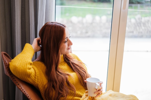 Jeune femme aux cheveux roux assis près de la fenêtre se détendre tout en prenant un café chaud Concept de vie détente tranquillité