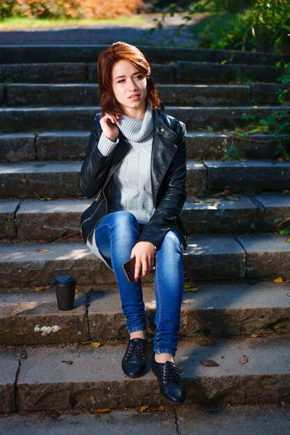 Une jeune femme aux cheveux roux dans une veste en cuir et un jean est assis sur les marches avec une tasse de café