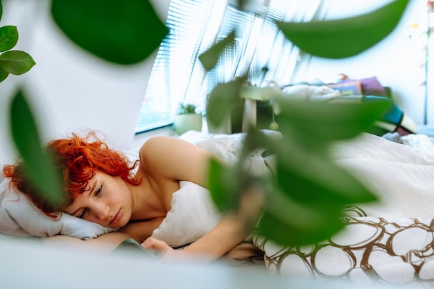 Une jeune femme aux cheveux roux est allongée dans le lit sous une couverture et utilise un smartphone pour les vacances du matin.