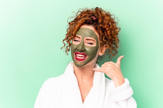 Jeune femme aux cheveux roux avec un masque facial à l'aloe vera après un bain isolé montrant un geste d'appel de téléphone portable avec les doigts