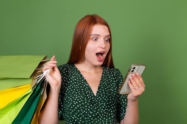 Jeune femme aux cheveux roux sur un mur vert avec des sacs à provisions et un téléphone portable choqué étonné