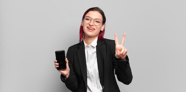 Jeune femme aux cheveux roux souriante et à la recherche heureuse, insouciante et positive, gesticulant la victoire ou la paix d'une seule main. concept d'entreprise