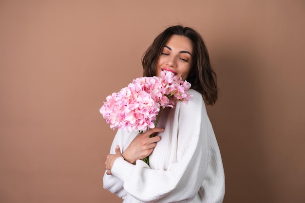 Une jeune femme aux cheveux volumineux ondulés sur fond beige avec un brillant à lèvres rouge à lèvres rose vif dans un pull blanc tient un bouquet de fleurs roses