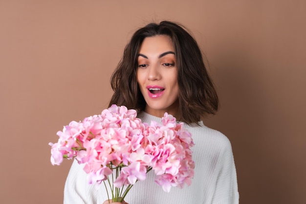 Photo une jeune femme aux cheveux volumineux ondulés sur fond beige avec un brillant à lèvres rouge à lèvres rose vif dans un pull blanc tient un bouquet de fleurs roses