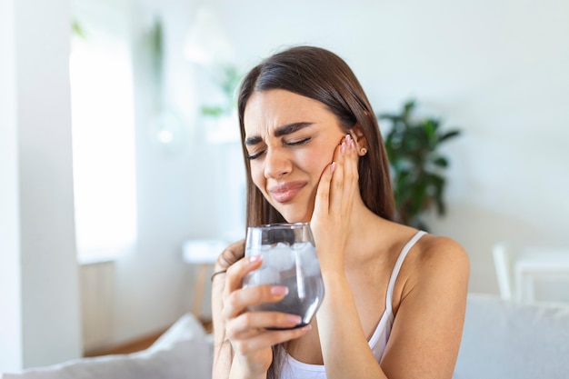 Jeune femme aux dents sensibles faisant mal et tenant un verre d'eau froide avec de la glace à la maison