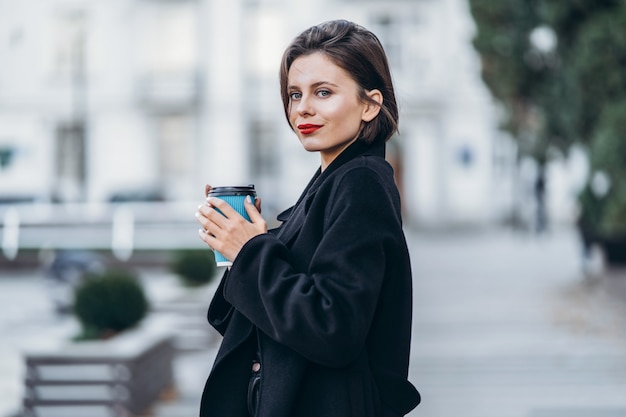 Jeune femme aux lèvres rouges et coupe courte à l'extérieur tenant une tasse de café dans ses mains