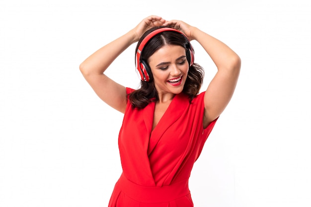 Une jeune femme aux lèvres rouges, maquillage lumineux, cheveux longs ondulés sombres, dans un costume rouge, des lunettes noires avec des piles transparentes se tient et écoute de la musique dans les écouteurs et les danses