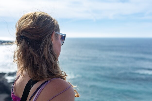 Jeune femme aux longs cheveux blonds assis sur la plage et regardant l'horizon. Océan bleu et rivage rocheux.