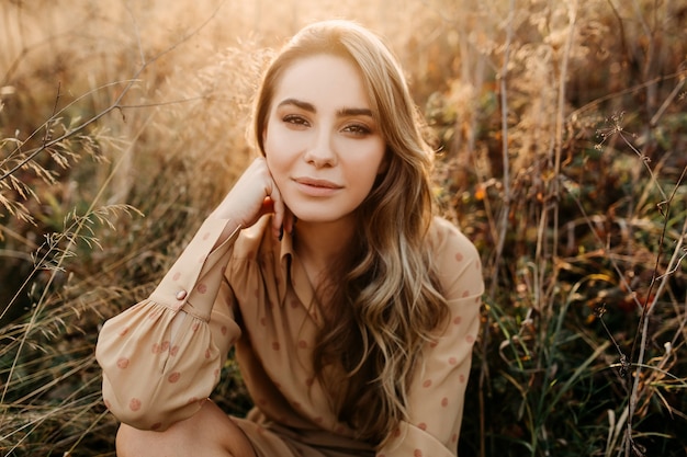 Jeune femme aux longs cheveux ondulés blonds souriant assis dans un champ avec de grandes herbes sèches