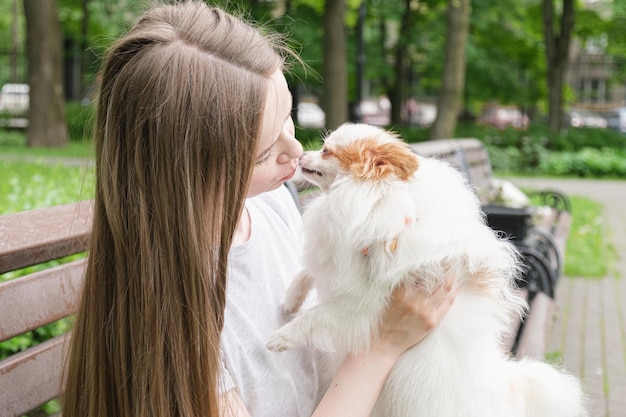 Une jeune femme aux longs cheveux raides est assise sur un banc dans un parc et embrasse un petit chien moelleux dans ses bras