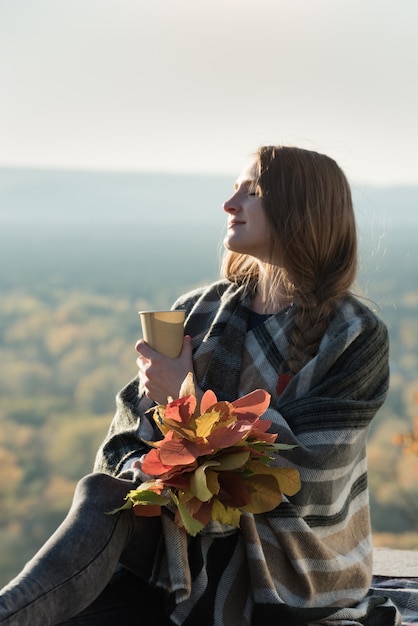 Jeune femme aux yeux fermés est assise sur une colline. Gobelet en papier dans les mains et un bouquet de feuilles jaunes. Profiter d'une journée ensoleillée
