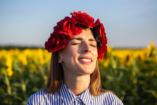 Jeune femme aux yeux fermés portant une couronne de fleurs rouges sur un champ de tournesol au coucher du soleil.