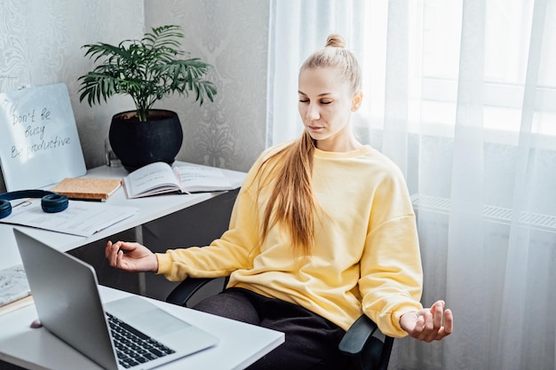 Jeune femme aux yeux fermés près d'un ordinateur portable au bureau à domicile pratiquant la méditation à table pigiste