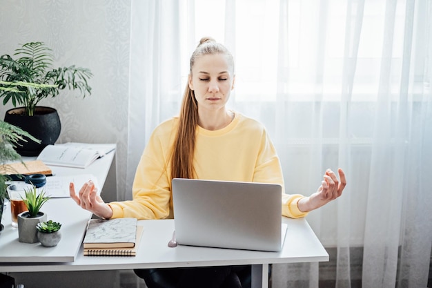 Jeune femme aux yeux fermés près d'un ordinateur portable au bureau à domicile pratiquant la méditation à table pigiste