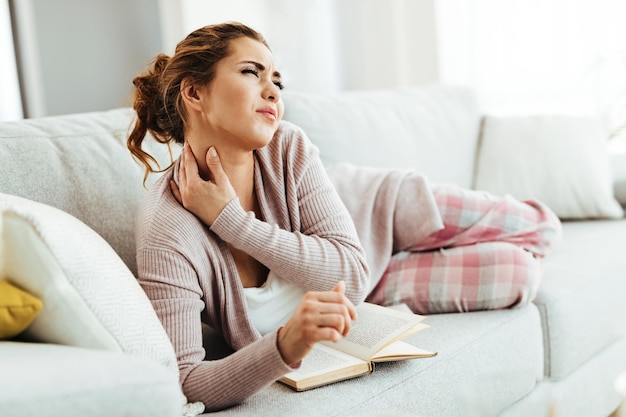 Jeune femme ayant mal au cou allongée sur un canapé et lisant un livre dans le salon