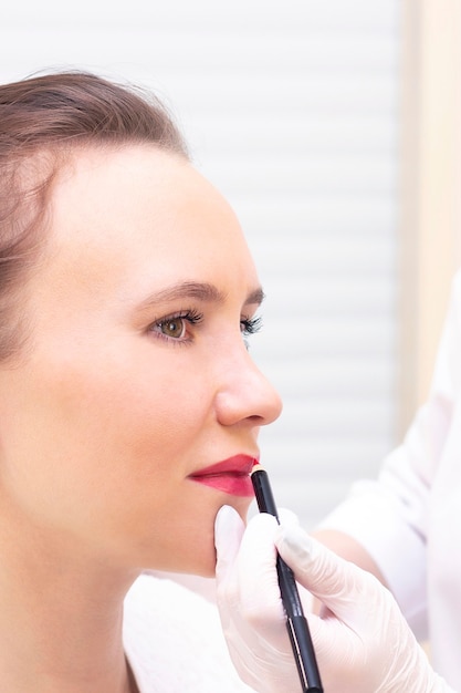 Jeune femme ayant un maquillage permanent sur ses lèvres au salon de l'esthéticienne. Maquillage permanent (tatouage). dessiner un contour avec un crayon à lèvres blanc. photo verticale
