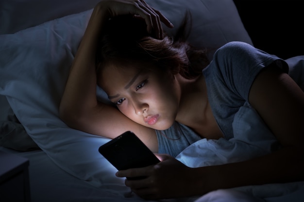 Jeune femme ayant les yeux endoloris et fatigués lors de l'utilisation de smartphone en position couchée dans son lit la nuit