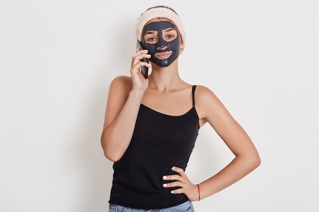 Jeune femme avec bandeau sur la tête et masque d'argile du visage noir, parler par téléphone. Traitements de beauté Spa, soins de la peau à la maison, femme heureuse contre le mur blanc.