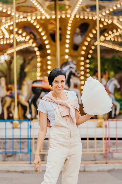 Jeune femme avec barbe à papa devant le carrousel avec éclairage nocturne au parc d'attractions.