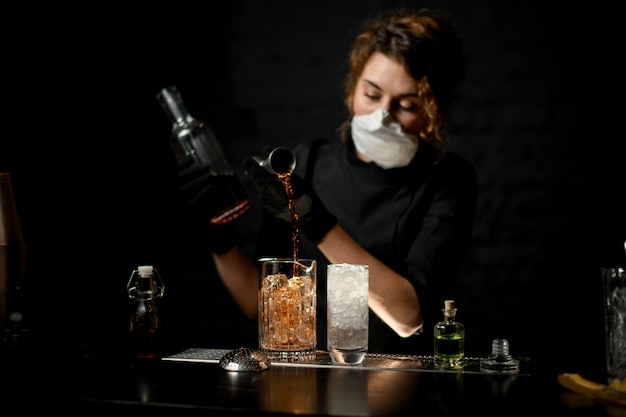 Jeune femme barman en masque médical verse une boisson alcoolisée à dark bar