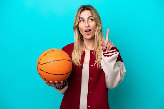 Jeune femme de basket-ball caucasienne isolée sur fond bleu pensant à une idée pointant le doigt vers le haut