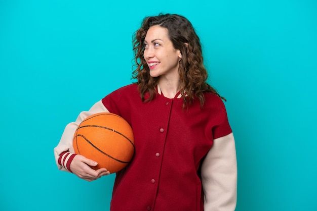 Jeune femme de basket-ball caucasienne isolée sur fond bleu regardant sur le côté et souriant