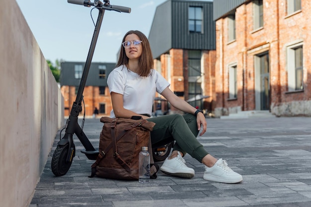 Jeune femme en baskets blanches est assise sur un scooter électrique sur fond de rue urbaine