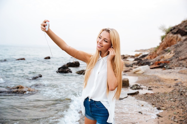 Jeune femme belle souriante écoutant de la musique et faisant selfie en se tenant debout sur la plage rocheuse