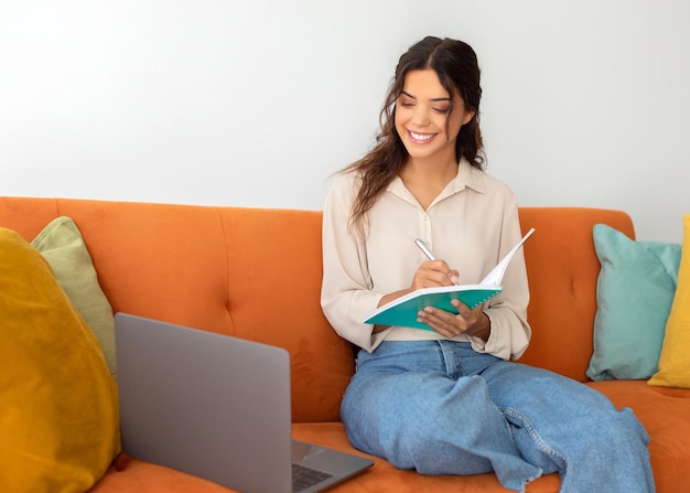 Une jeune femme belle et souriante prend des notes en étudiant avec un ordinateur portable à la maison