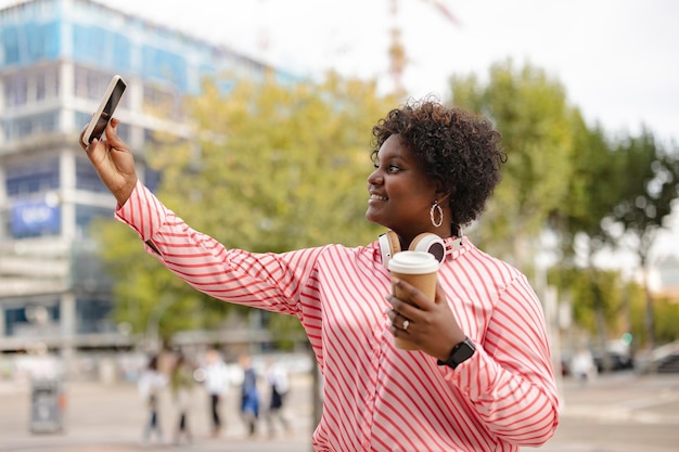 Jeune femme bien roulée afro-américaine buvant un café jetable et prend un selfie avec le smartphone dans la rue