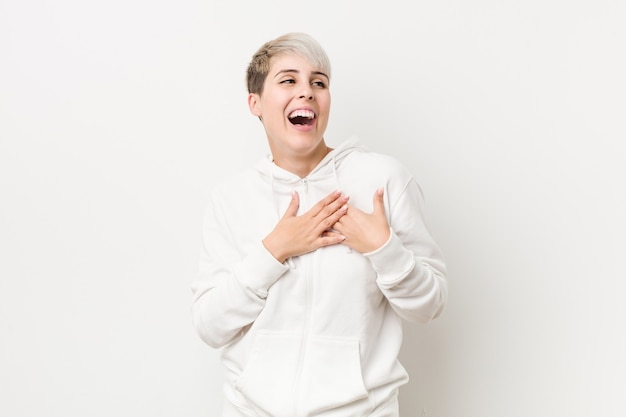 Jeune femme bien roulée portant un sweat à capuche blanc en riant en gardant les mains sur le cœur, le concept du bonheur.