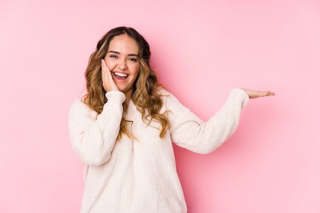 Jeune femme bien roulée posant dans un mur rose isolé détient un espace de copie sur une paume, gardez la main sur la joue. Etonné et ravi.