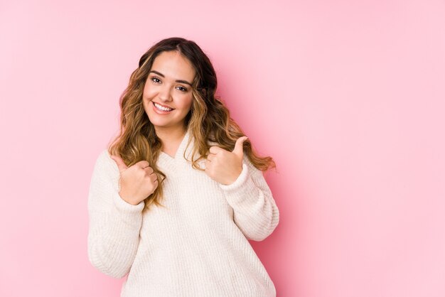 Jeune femme bien roulée posant dans un mur rose isolé levant les deux pouces vers le haut, souriant et confiant.