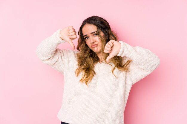 Jeune femme bien roulée posant dans un mur rose isolé montrant le pouce vers le bas et exprimant l'aversion.