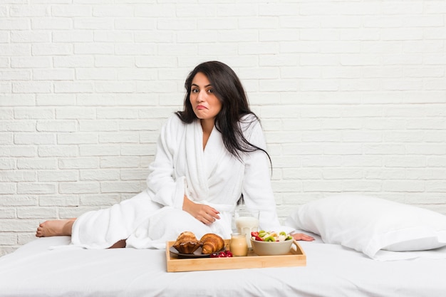 Jeune femme bien roulée prenant un petit déjeuner sur le lit hausse les épaules et les yeux ouverts confus.