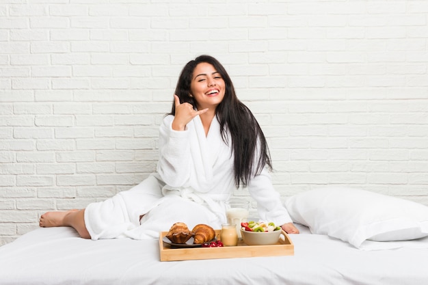 Jeune femme bien roulée prenant un petit déjeuner sur le lit montrant un geste d'appel avec les doigts.