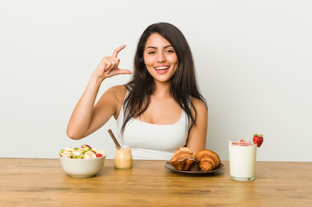 Jeune femme bien roulée prenant un petit déjeuner tenant quelque chose de peu avec les index, souriant et confiant.