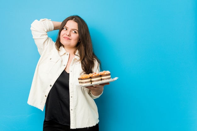 Jeune femme bien roulée tenant un cupcakes touchant l'arrière de la tête, pensant et faisant un choix.