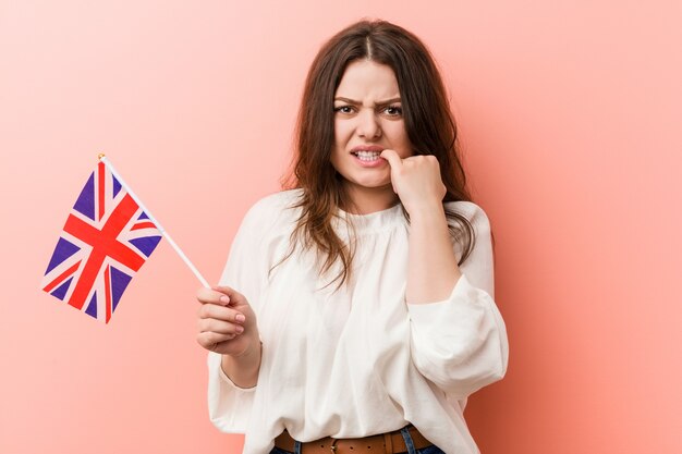 Jeune femme bien taille, curvy, tenant un drapeau du royaume-uni se ronger les ongles, nerveuse et très anxieuse.