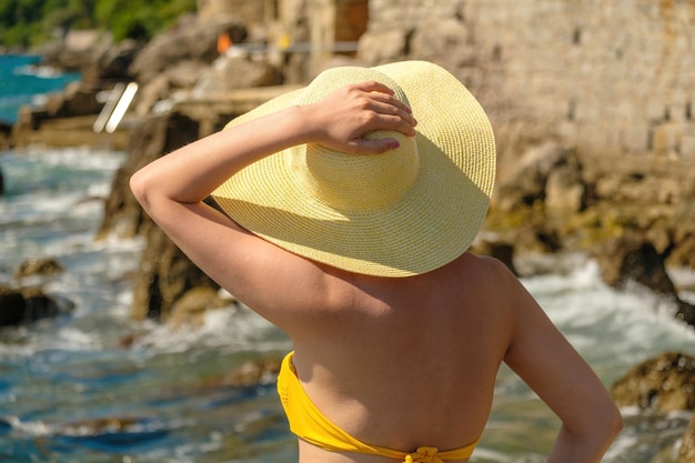 Jeune femme en bikini jaune tenant son chapeau et regardant la mer