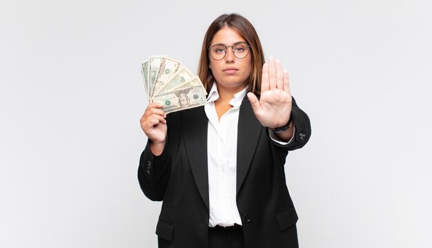 Jeune femme avec des billets à la sérieuse, sévère, mécontente et en colère montrant la paume ouverte faisant le geste d'arrêt