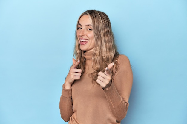 Une jeune femme blanche blonde dans un studio bleu pointant vers l'avant avec des doigts.